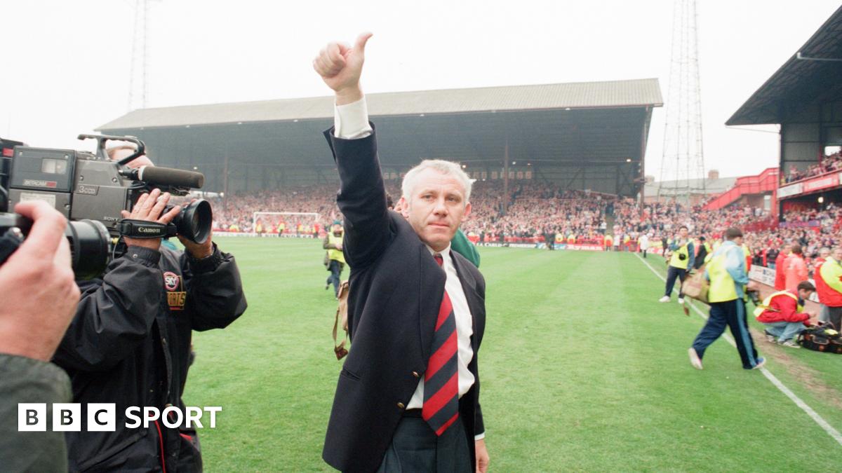 Peter Reid gives a thumbs-up gesture as he leaves the pitch at Roker Park following Sunderland's final game at the stadium. He has short grey hair and is wearing a dark blazer, white shirt and red tie. Two television cameramen are filming him. Thousands of fans are in the ground's stands.