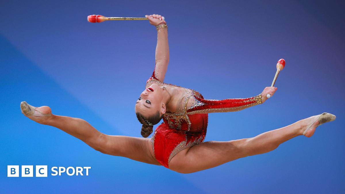 A rhythmic gymnast in a red leotard performs a mid-air split while holding two clubs against a blue gradient background.