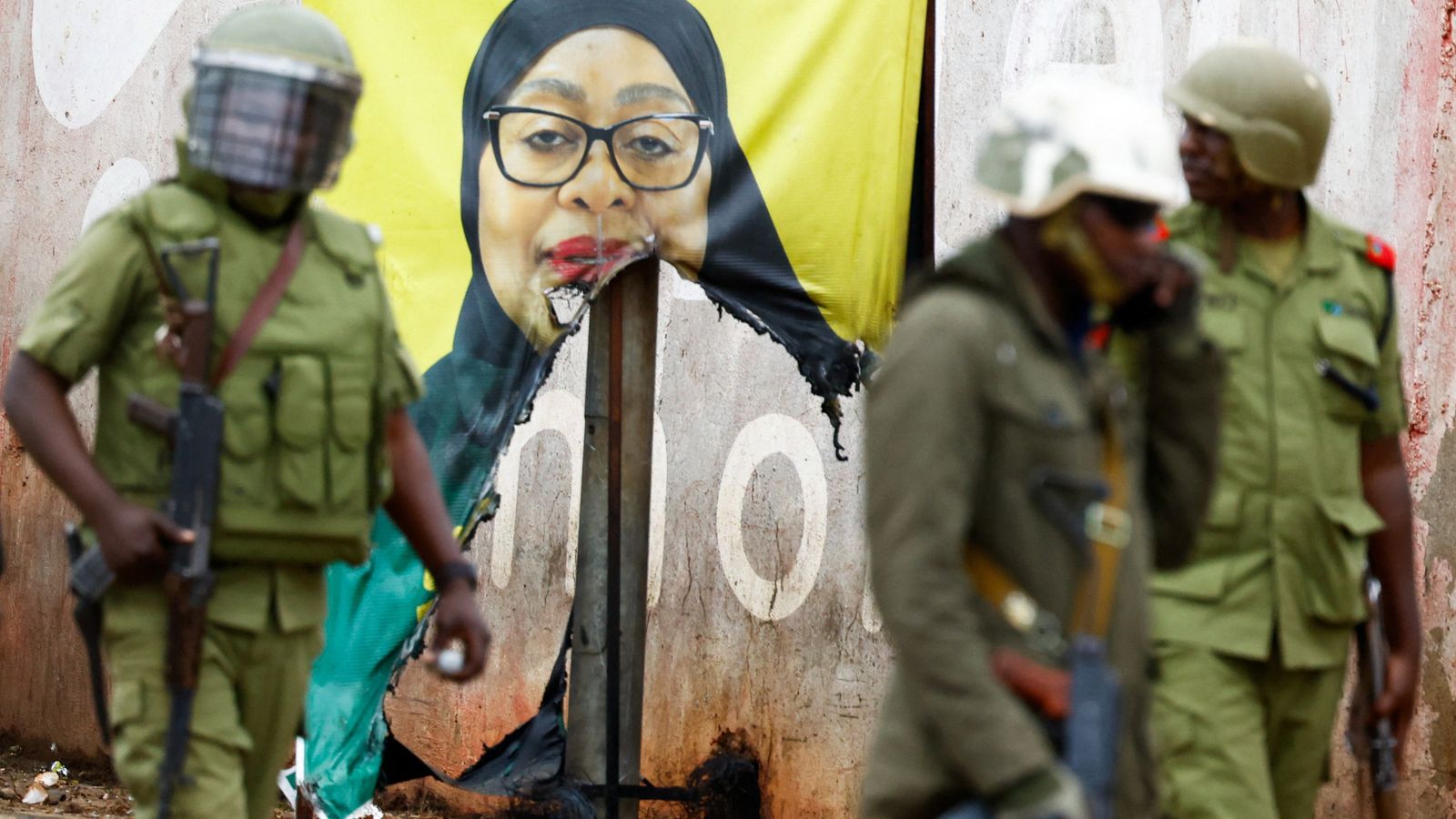 Tanzanian riot police officers walk past a vandalised campaign poster of President Samia Suluhu Hassan. Pic: Reuters