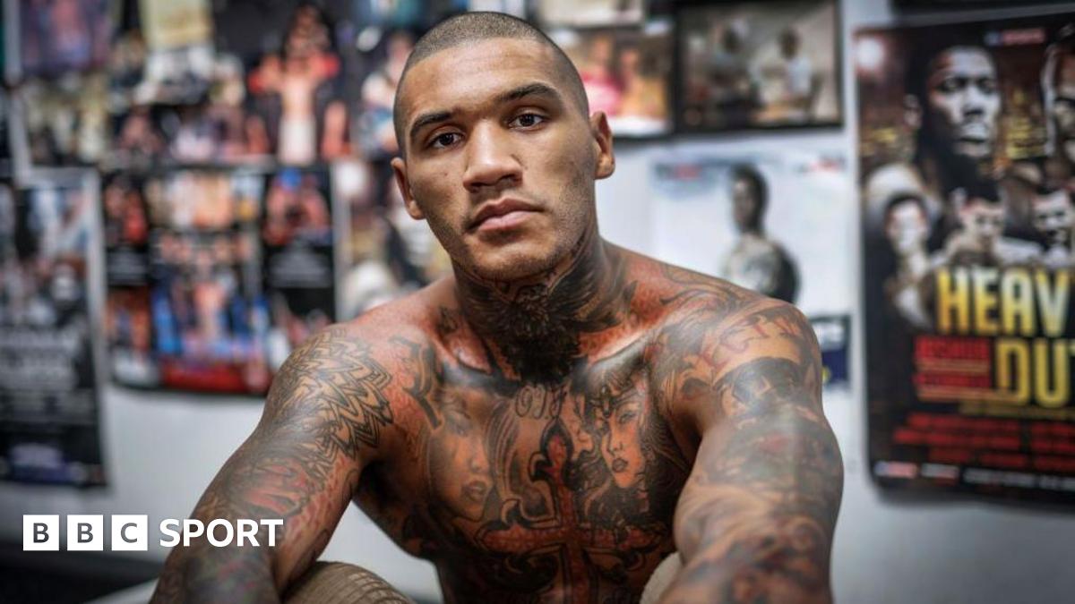Conor Benn is shirtless and sitting in front a wall of posters