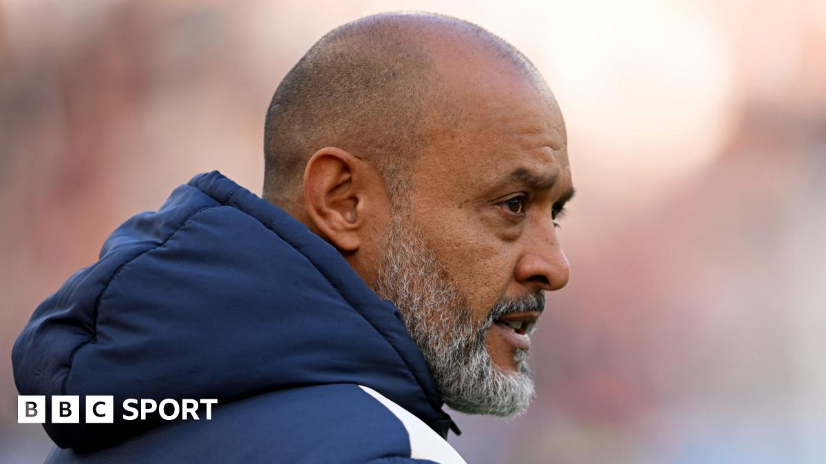 Nuno Espirito Santo, seen from the waist up, smiles during a promo shot for his appointment as the new West Ham United manager. He is wearing a blue West Ham tracksuit top with grey trim, is holding up a claret West Ham home shirt, and is standing in front of a sponsors board with many small rectangular logos on it, including the West Ham club crest